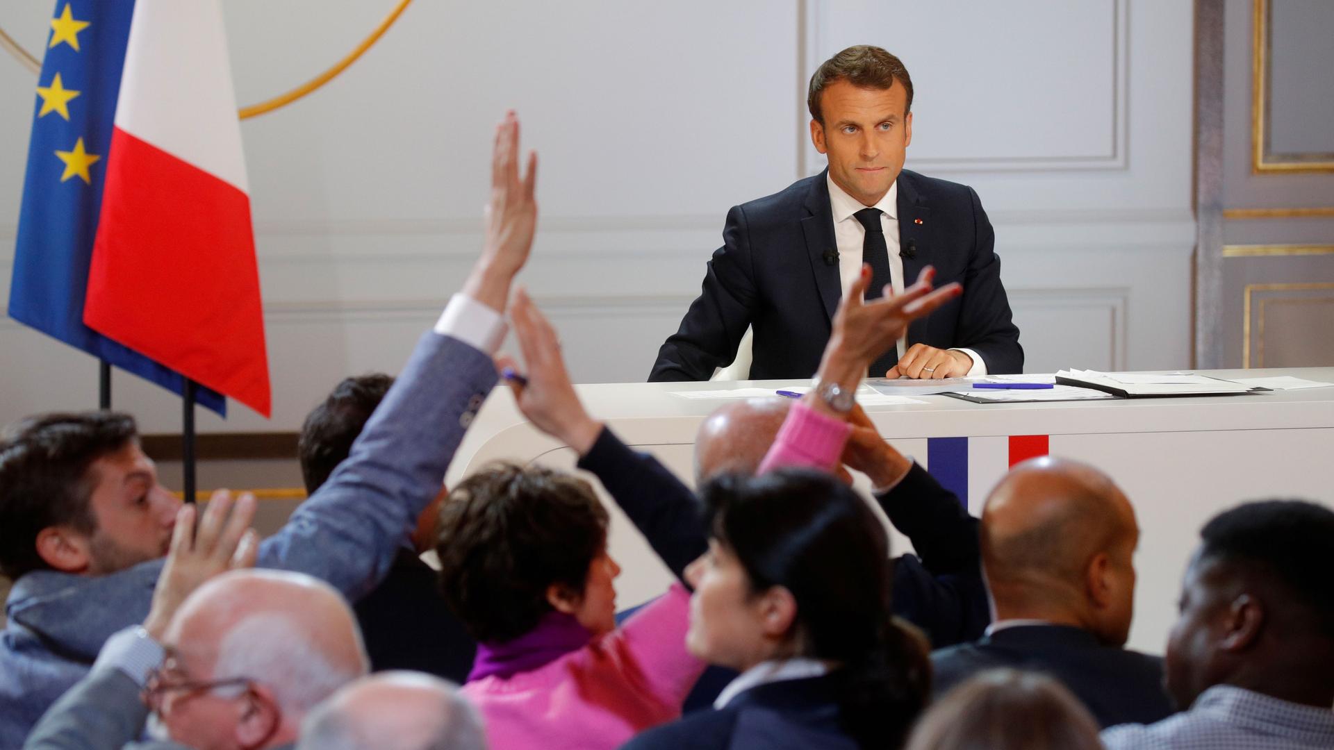Macron is at the front of a room and hands of journalists are in the air