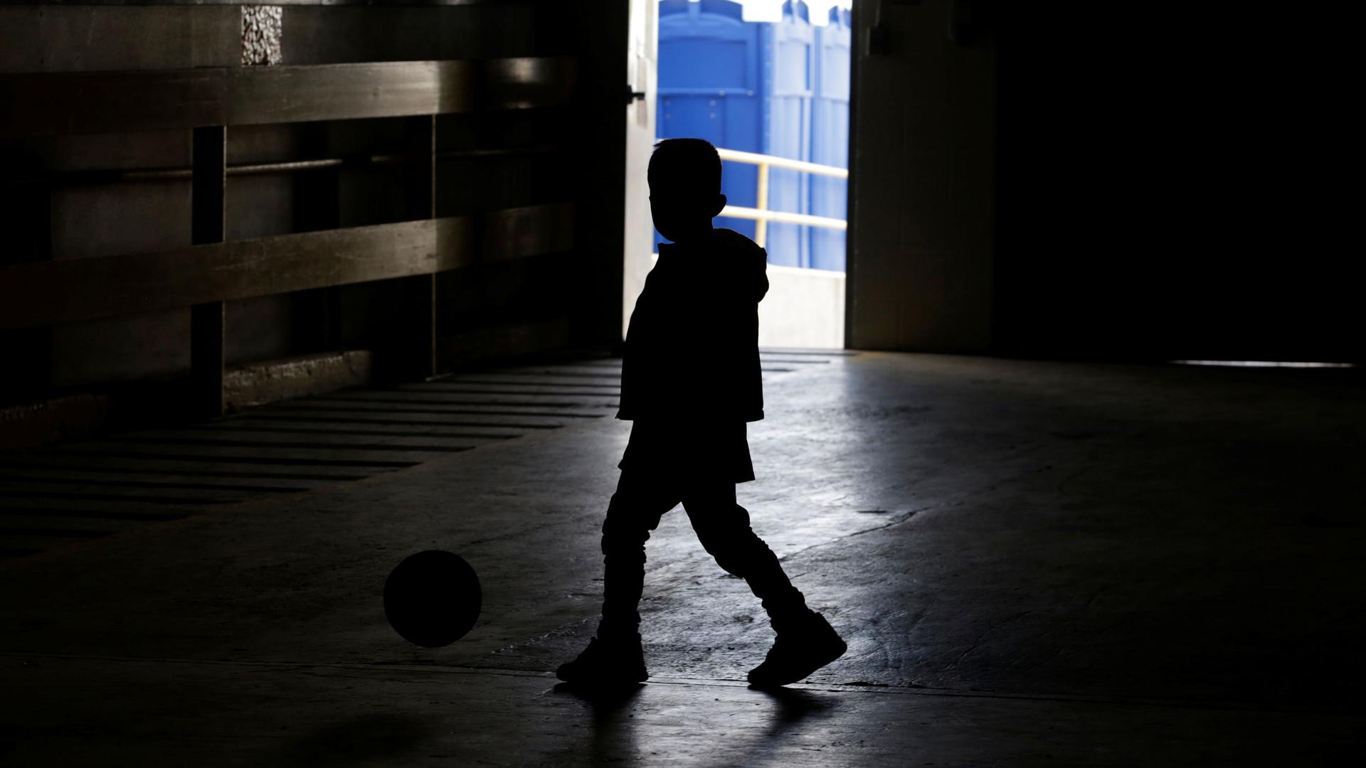 A child is shown in shadow playing with a soccer ball inside a temporary shelter.