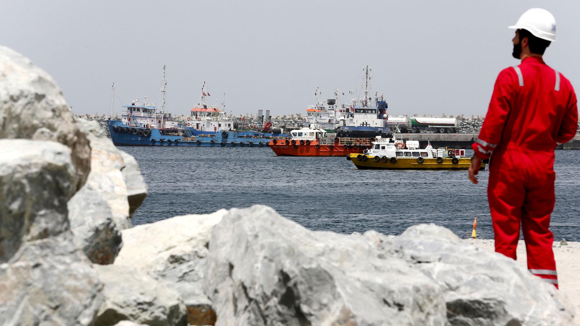 A port technical staff member is shown in the near ground wearing a red jumpsuit with several ships seen in the distance.
