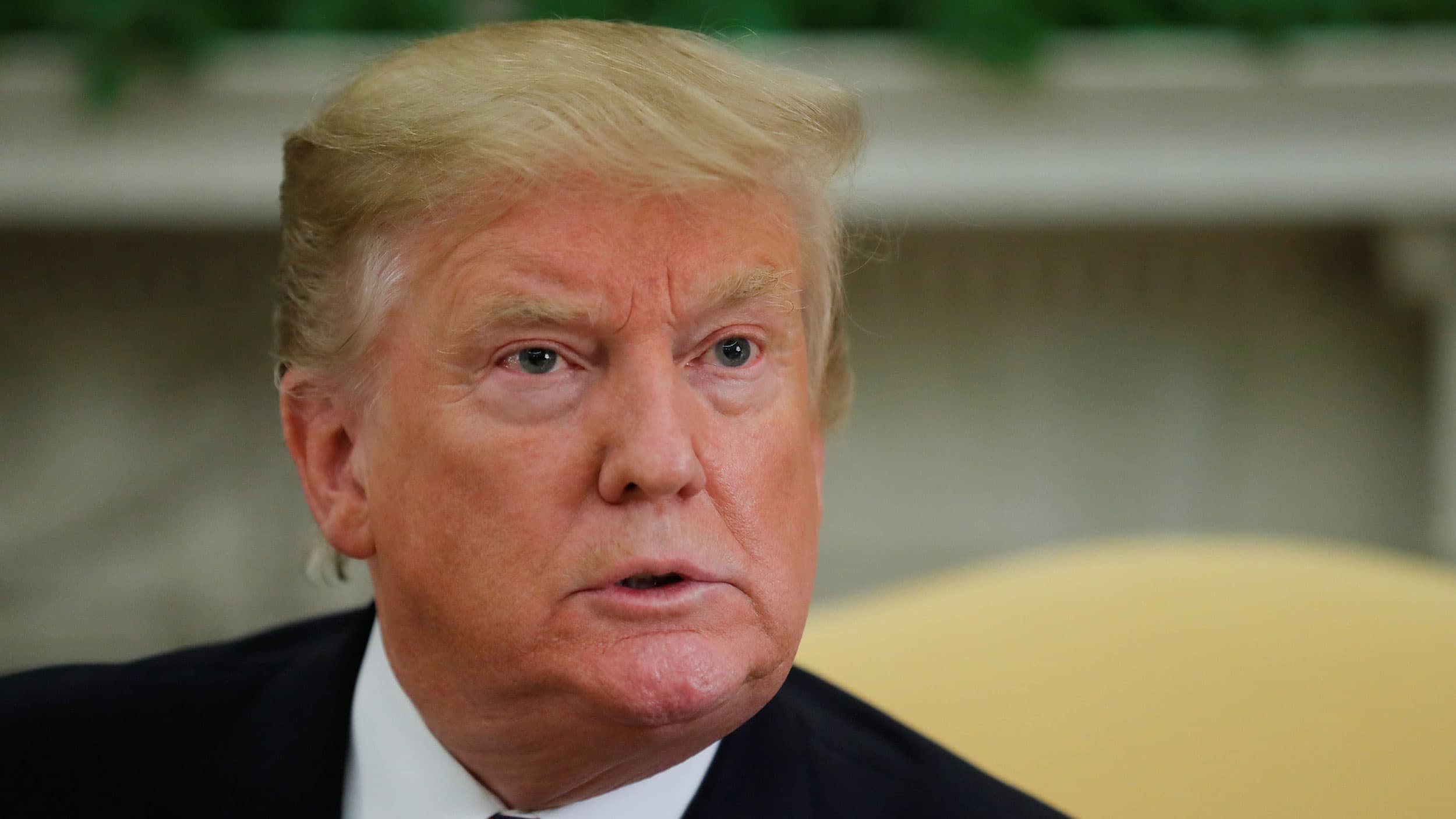 US President Donald Trump is shown in a close-up photograph listening to questions with a fixed gaze straight ahead.