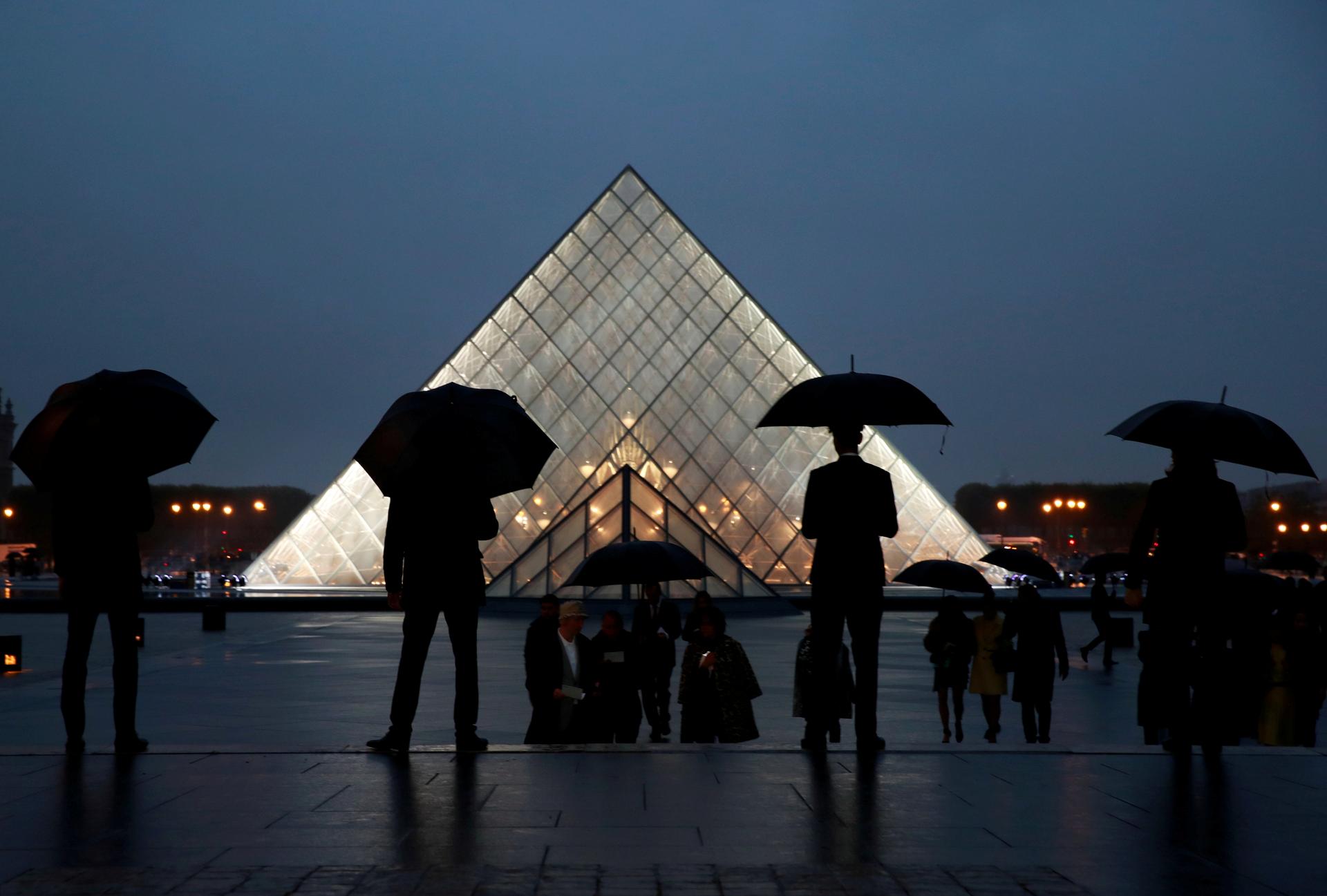 Silhouetted people with umbrellas stand in front of the Louvre pyramid at night.
