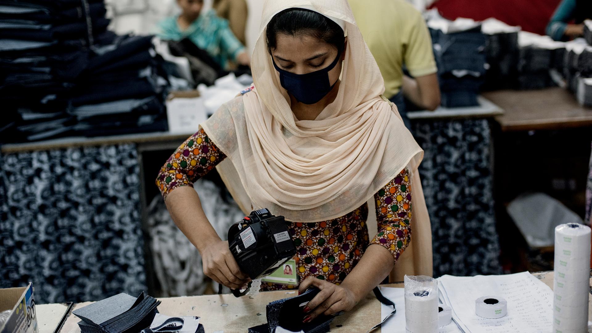 A garment factory employee, wearing a head scarf and face mask, working at a desk.