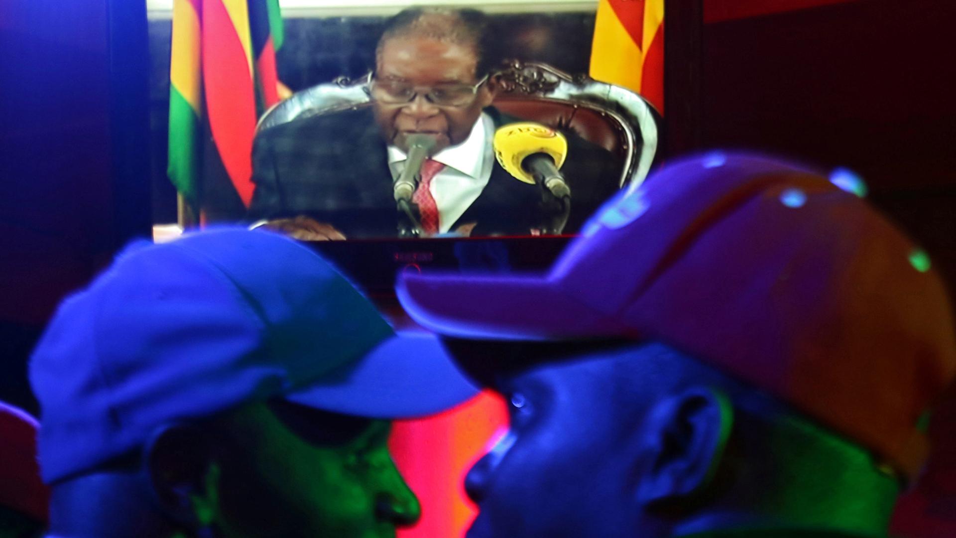 Two men in the foreground wearing baseball hats watch Zimbabwean President Robert Mugabe on a TV.