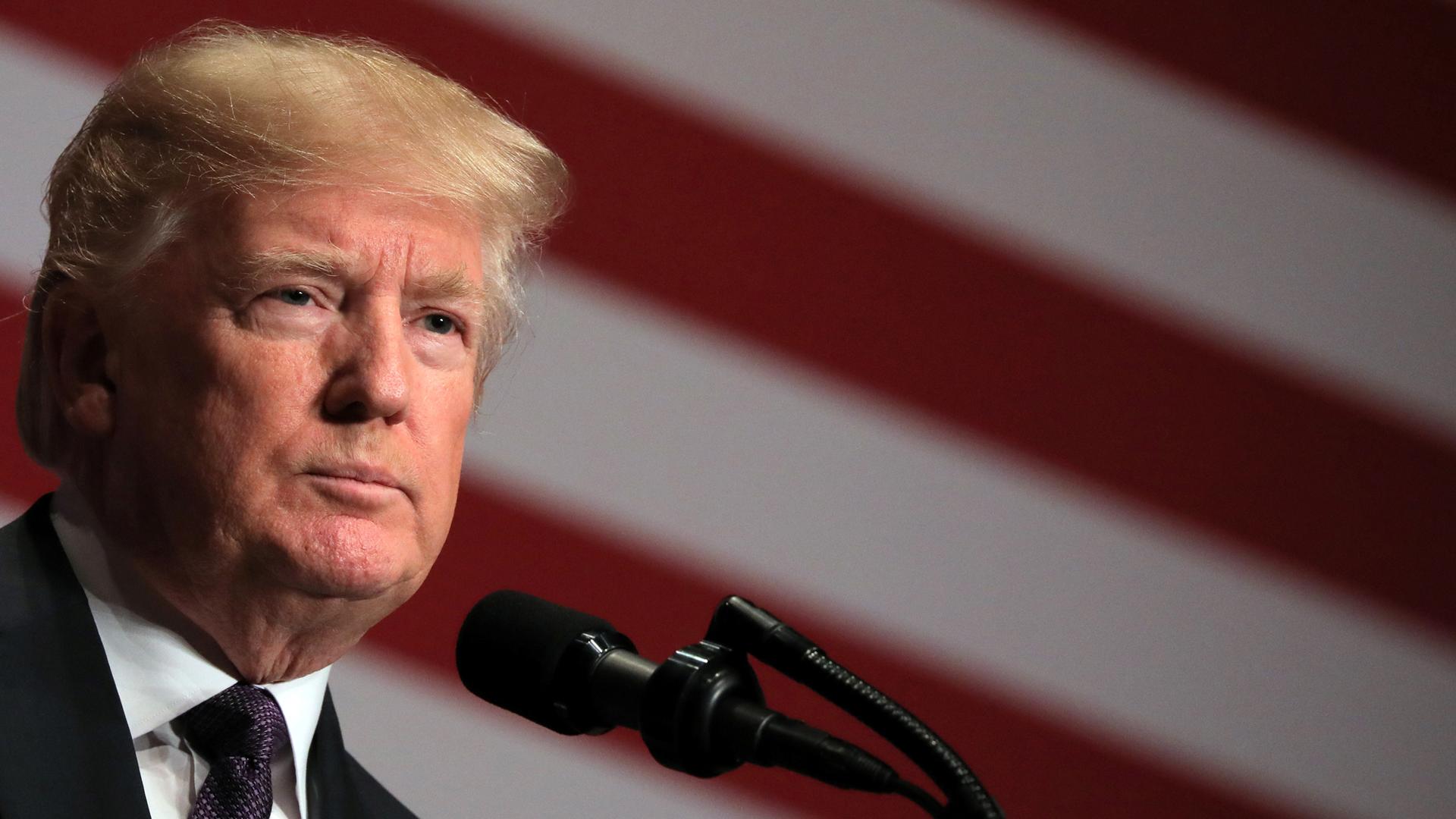 A close-up photo of President Donald Trump with a US flag blurred in the background.