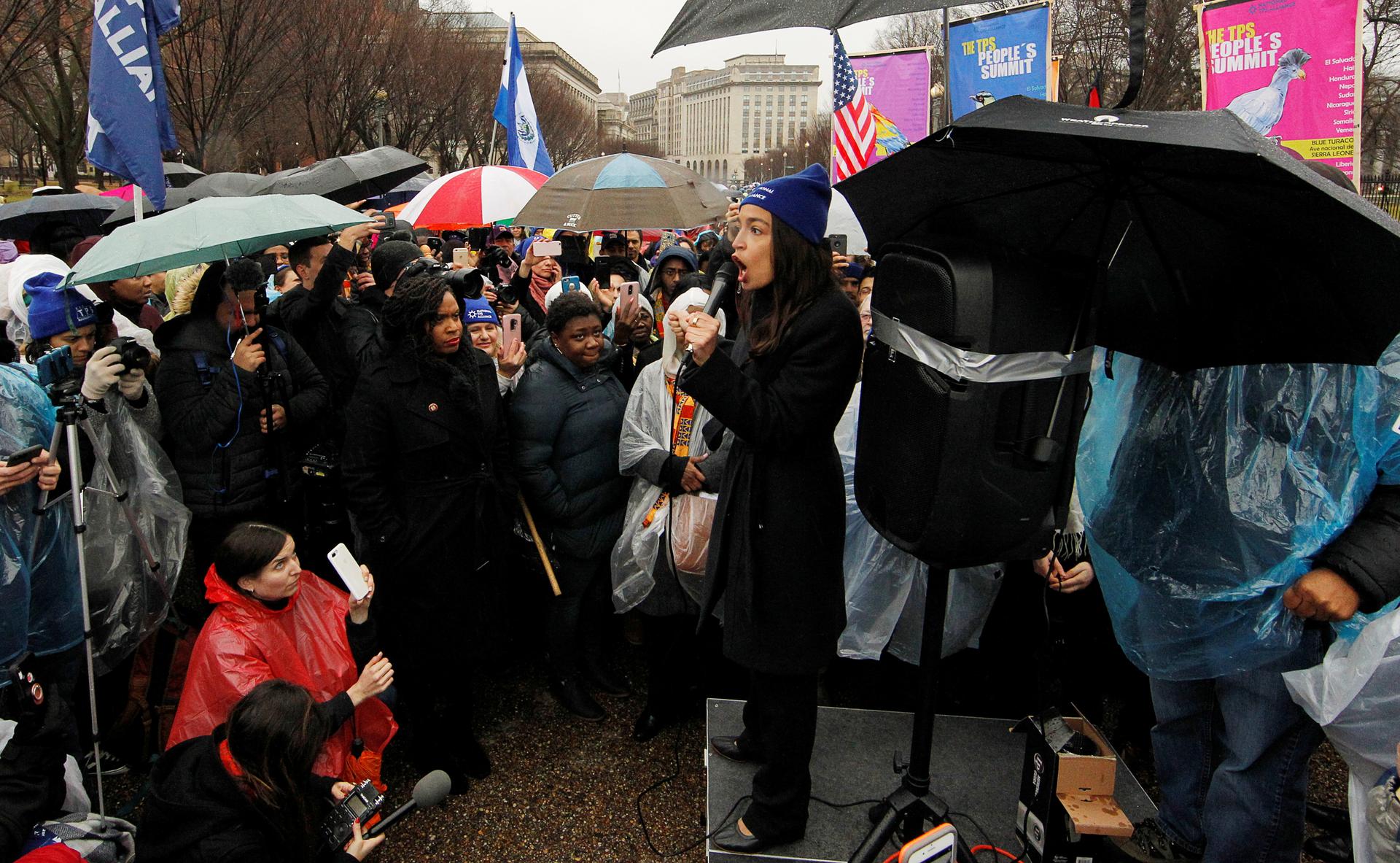 A woman with a microphone addresses the crowd surrounding her in the rain.