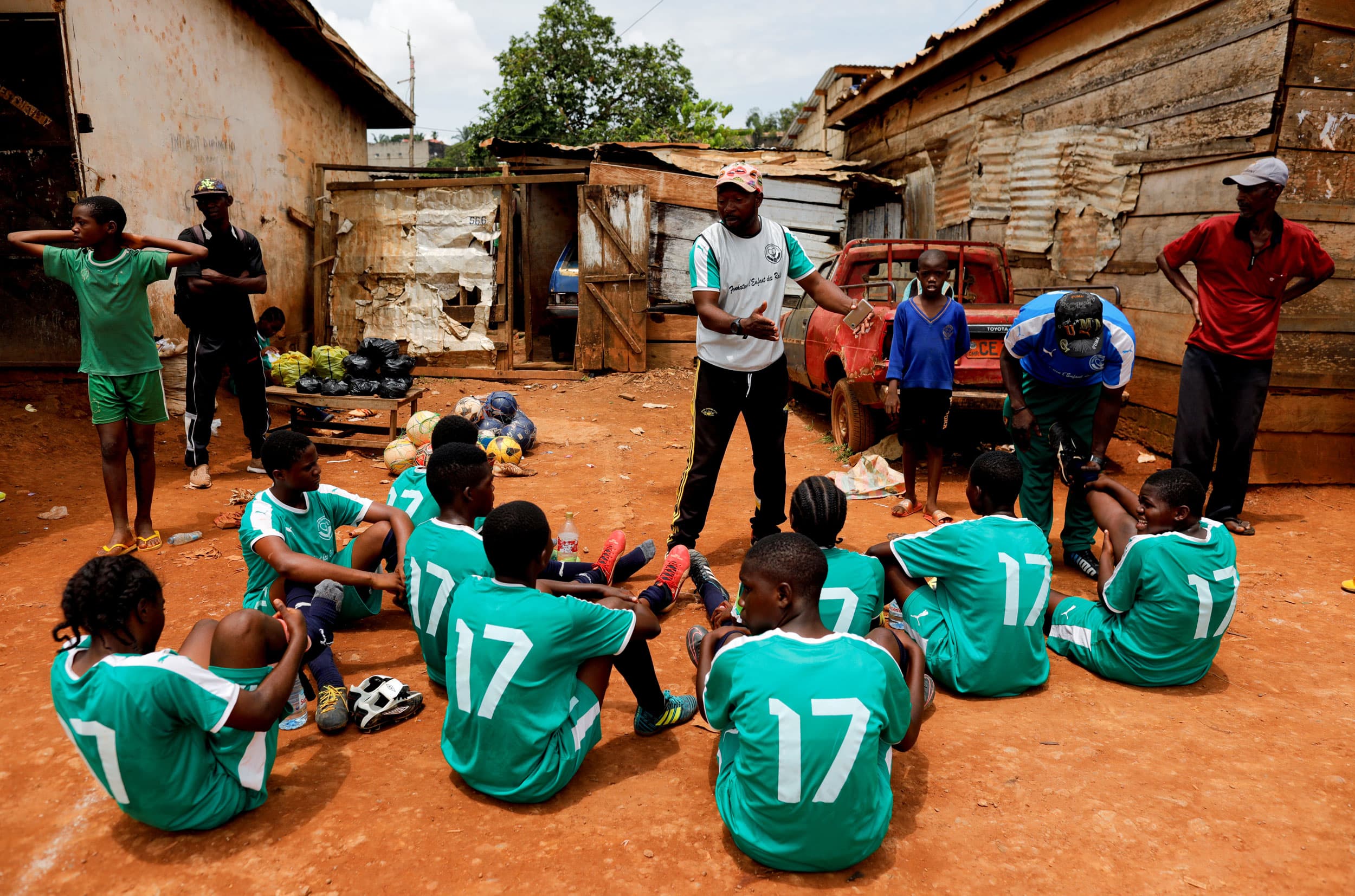 A coach is standing in front of his team whom are all sitting on the ground wearing green uniforms.