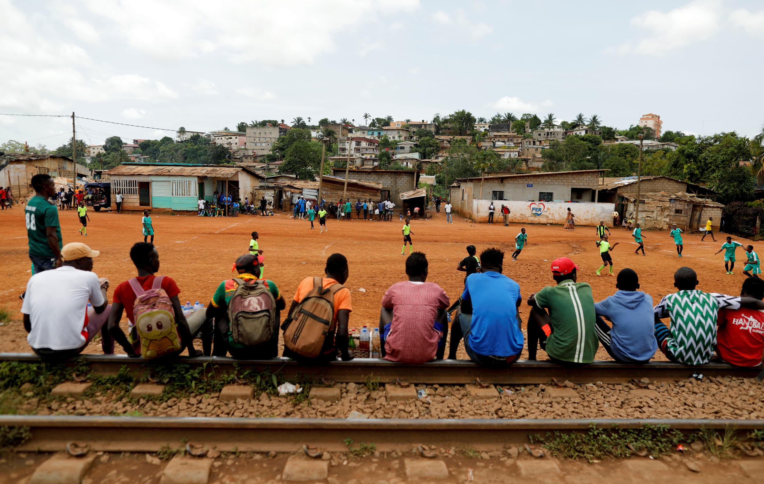 A row of people are shown sitting on railroad track with a soccer match taking place in the distance.