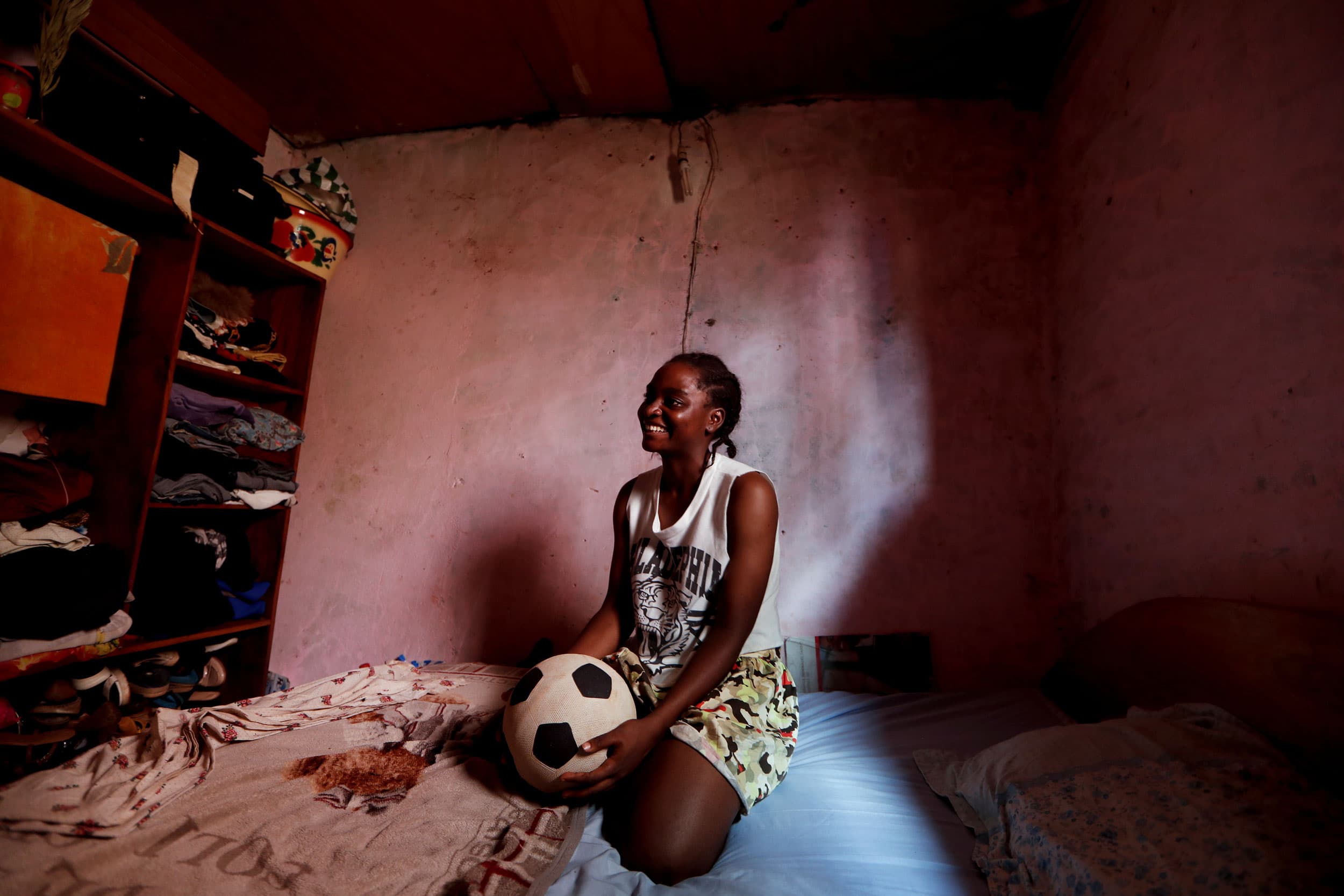 A young woman is shown sitting on her bed holding a soccer ball.