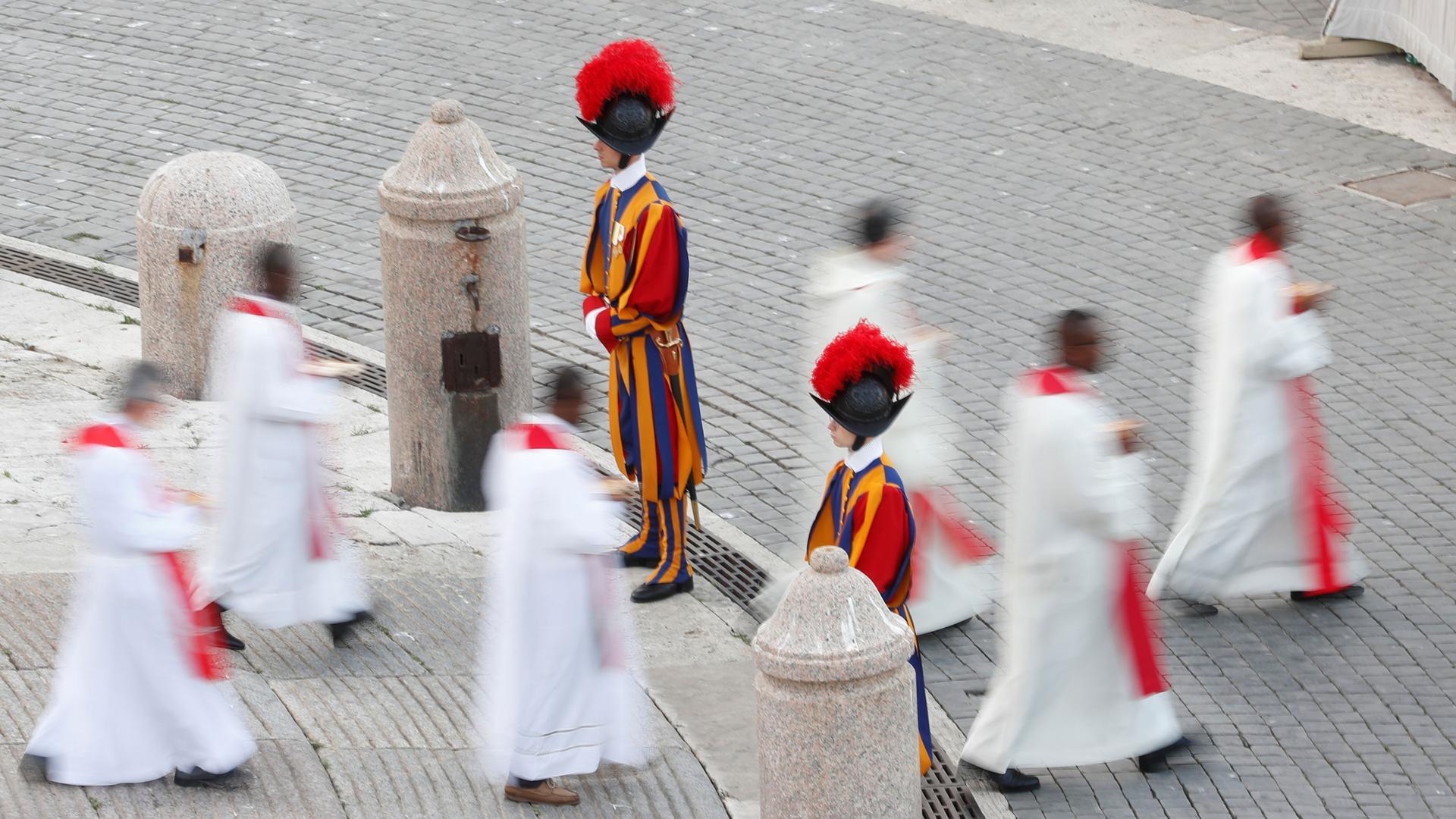 Several Catholic priests wearing white robes are shown walking past two guards in traditional striped uniforms.