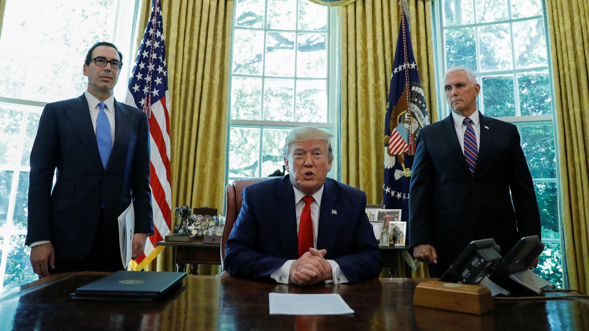 US President Donald Trump is shown sitting at a large wooden desk with his hands folded and wearing a blue suit and red tie.