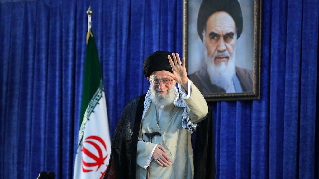 Iran's Supreme Leader Ayatollah Ali Khamenei is shown waving while standing next to an Iranian flag.