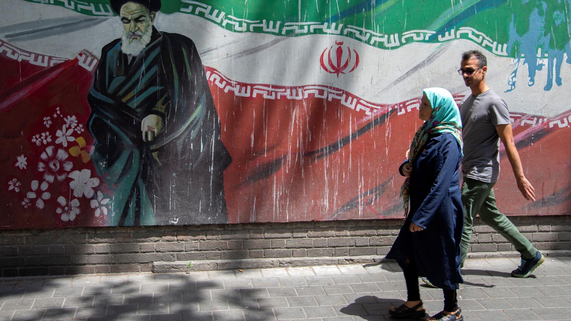 A woman wearing a shawl and a man wearing glasses are shown walking past a mural of Ayatollah Ruhollah Khomeini in front of an Iranian flag.