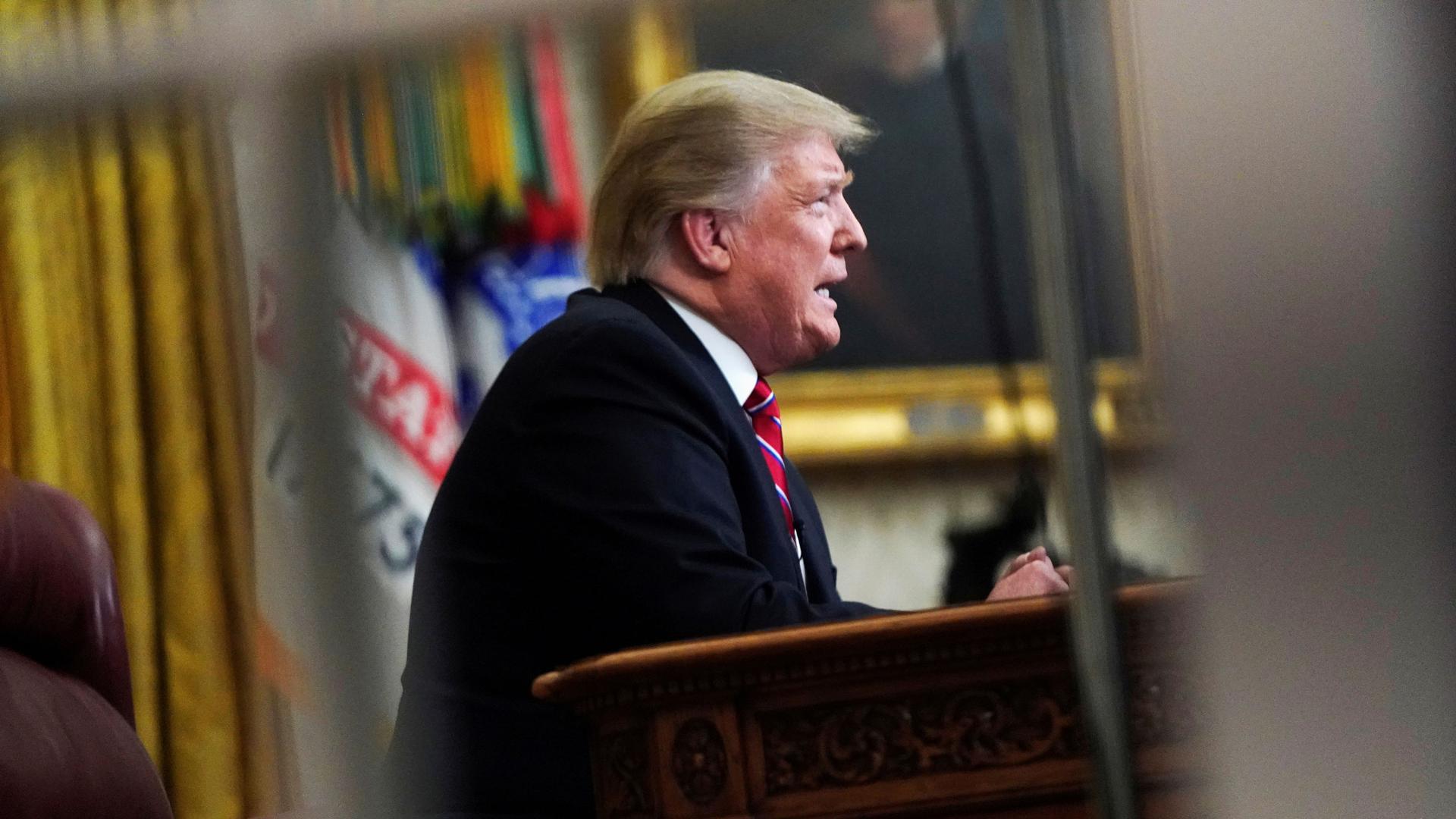 A photograph of President Donal Trump is seen through a window pane showing him sitting at a desk.