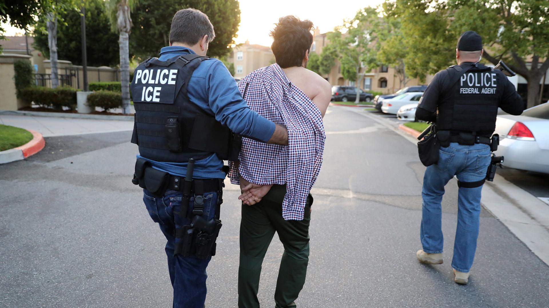 A man wearing a police vest reading POLICE ICE leads another man in handcuffs away. The handcuffed man has an open shirt draped over his shoulders. To the right is another man with a vest that says POLICE federal agent.