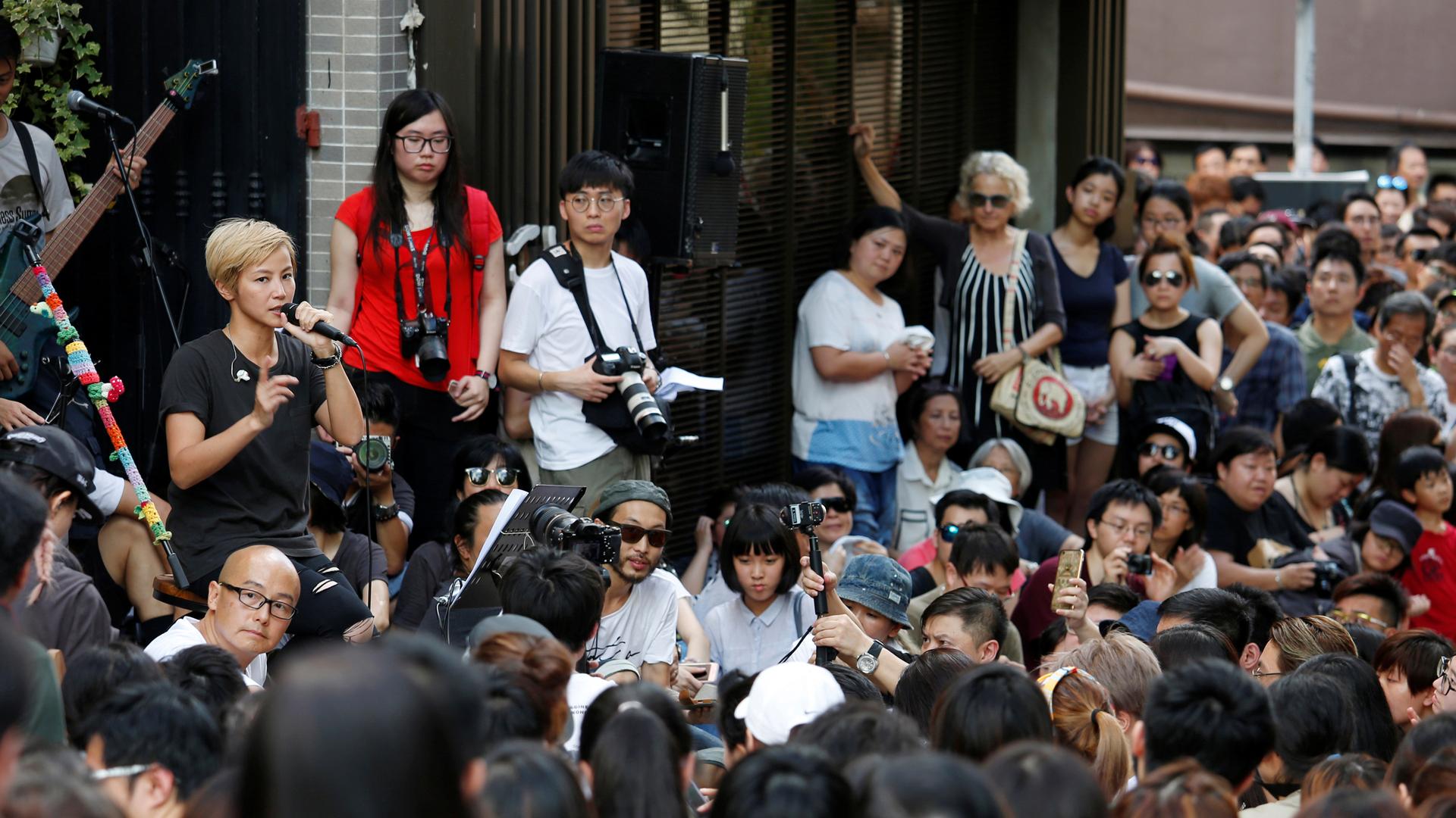 Thousands of people cram the streets of Hong Kong, walking off a freeway exit. The buildings of the city are behind them