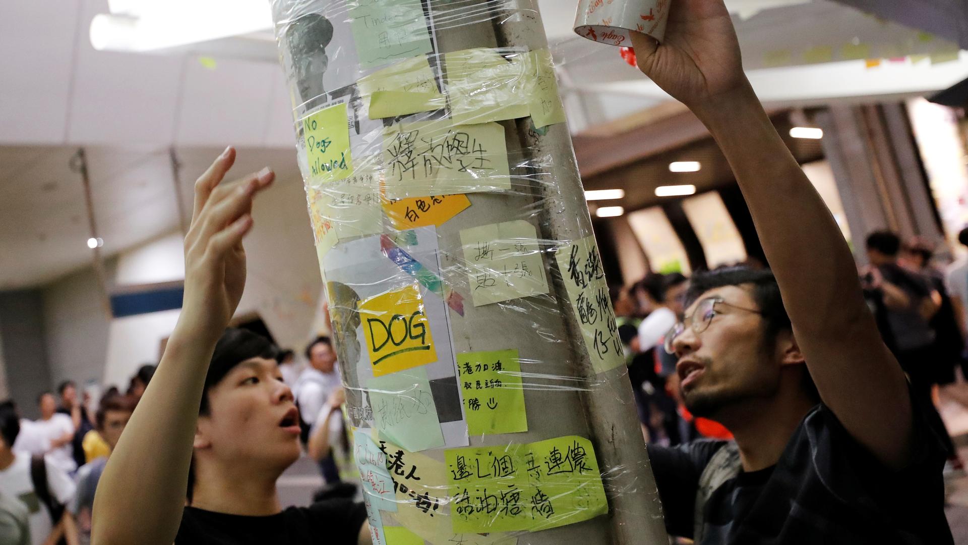 Two people wrap tape around a column covered in post-it notes to protect the "Lennon Wall"