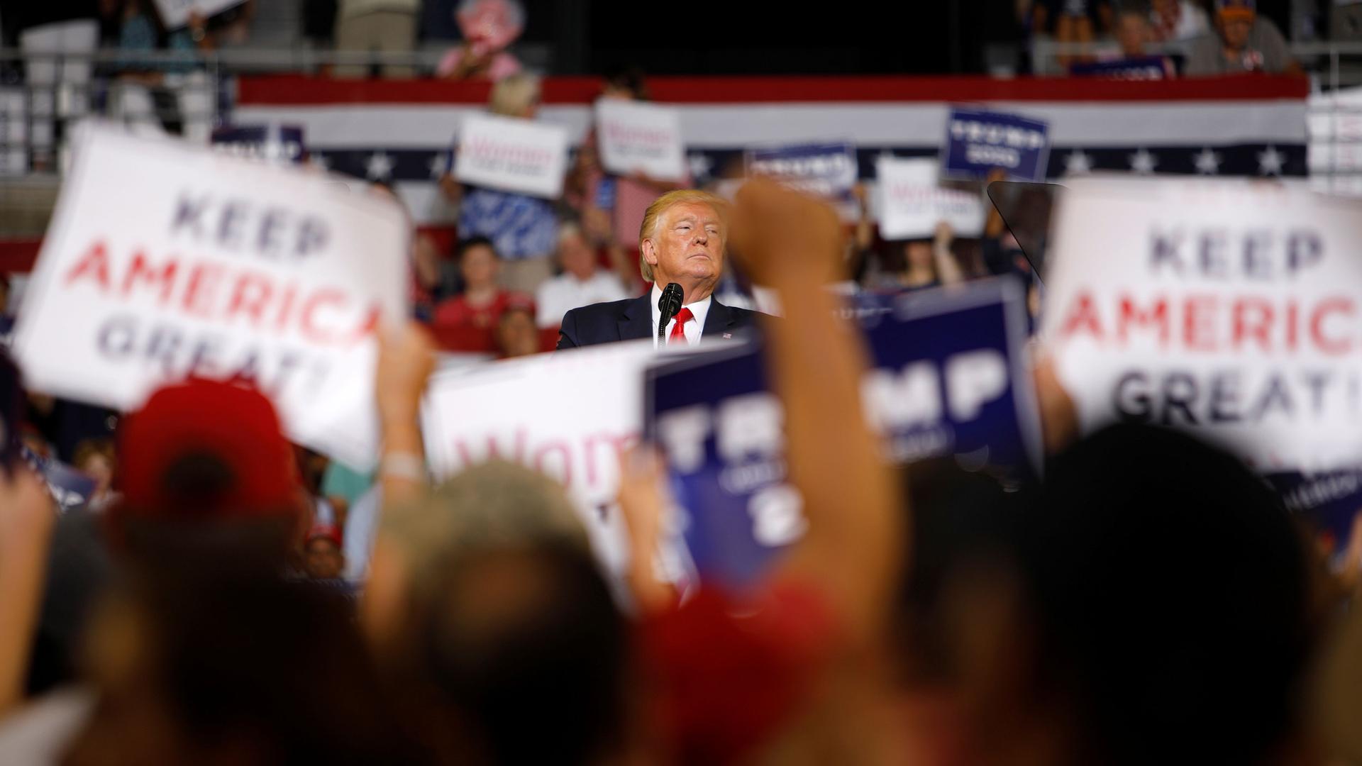 A large crowd of people are shown in soft focus holding placards with Donald Trump in center frame in focus.