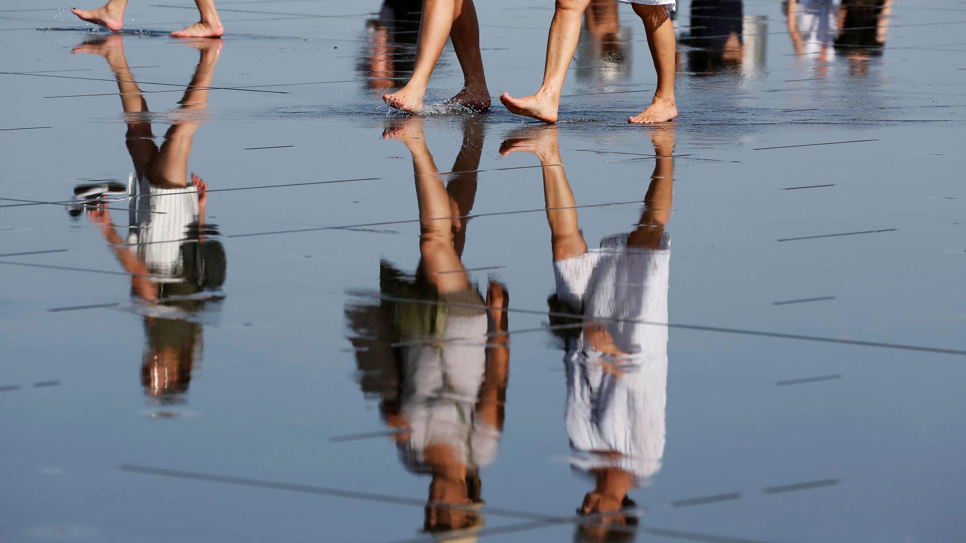 The reflection of several women are shown in water with just the bottom part of their legs shown at the top of the photograph.