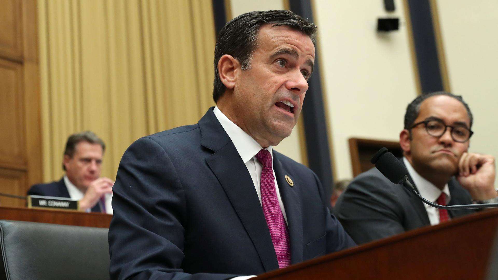 Rep. John Ratcliffe is shown speaking behind a microphone and wearing a blue suit coat with a pink tie.