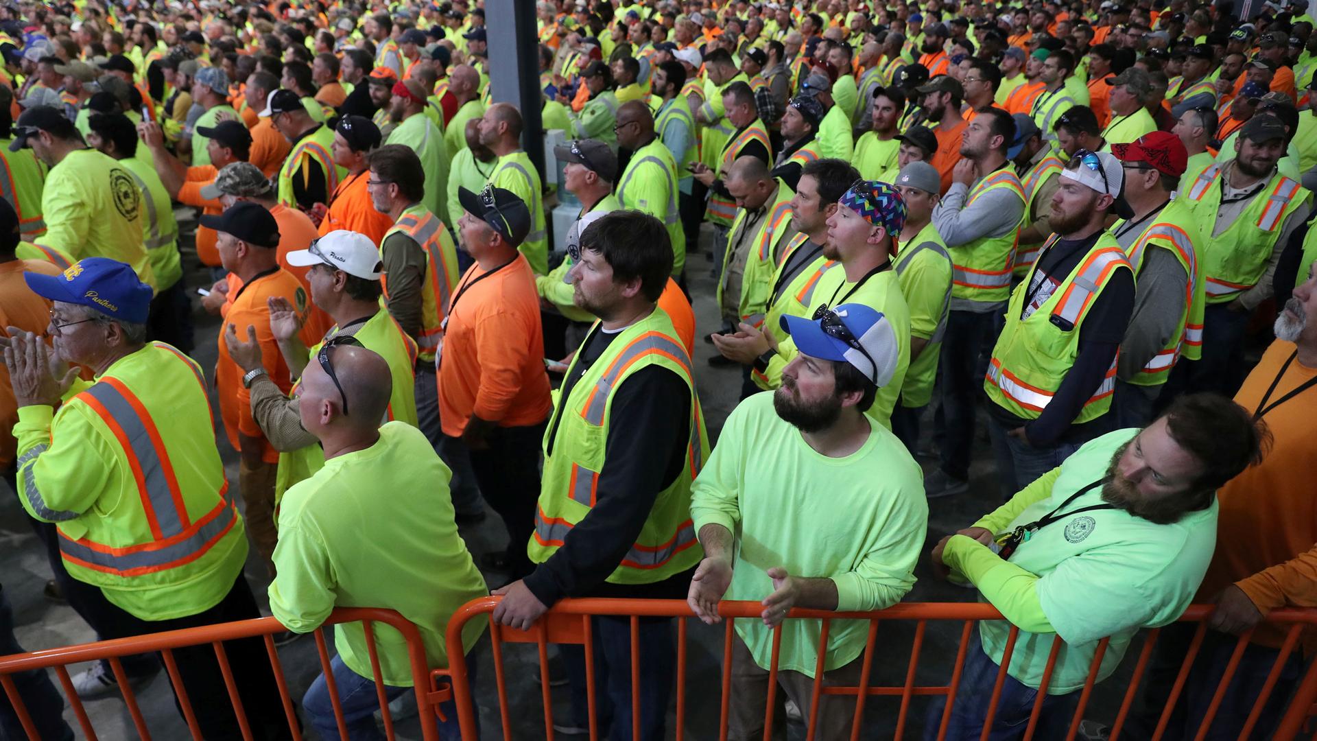 A crowd of workers in bright florescent colors stand and listen to a speaker