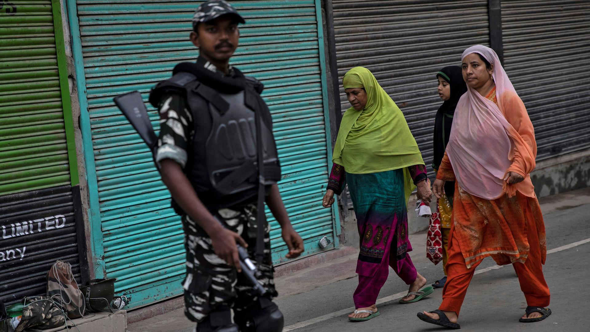 Three women are shown in colorful attire and head coverings walking past an armed security official.