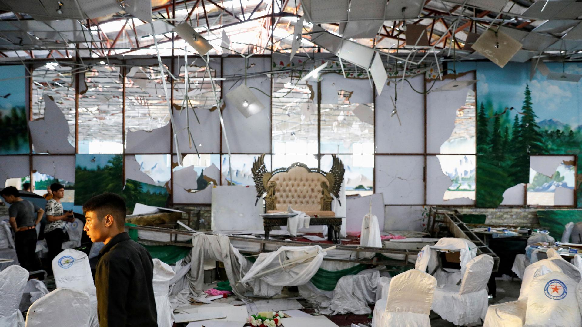 A young man is show walking off to the left through the debris room filled with white-covered chaies and a ceiling falling down.