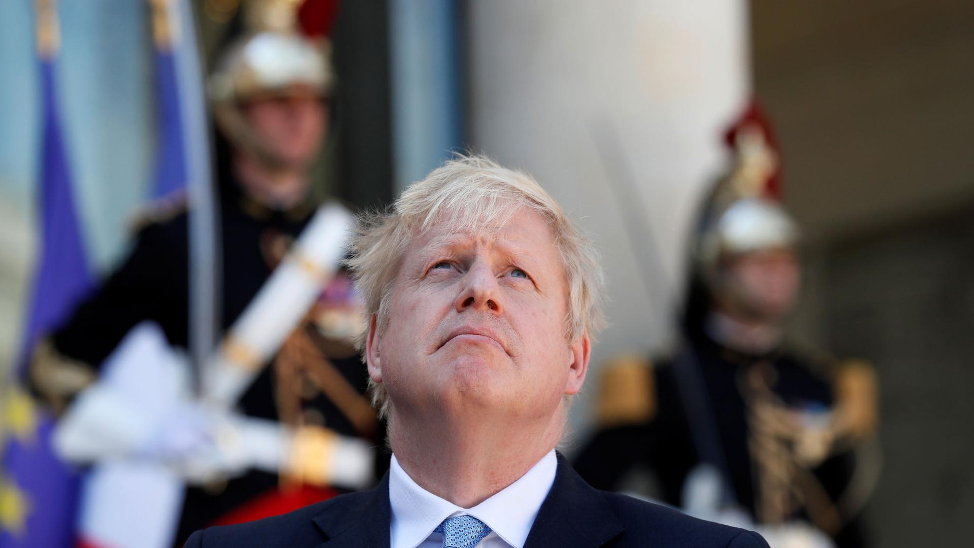 British Prime Minister Boris Johnson is shown looking up and wearing a suit and blue tie with Élysée Palace guards in soft focus in the background.