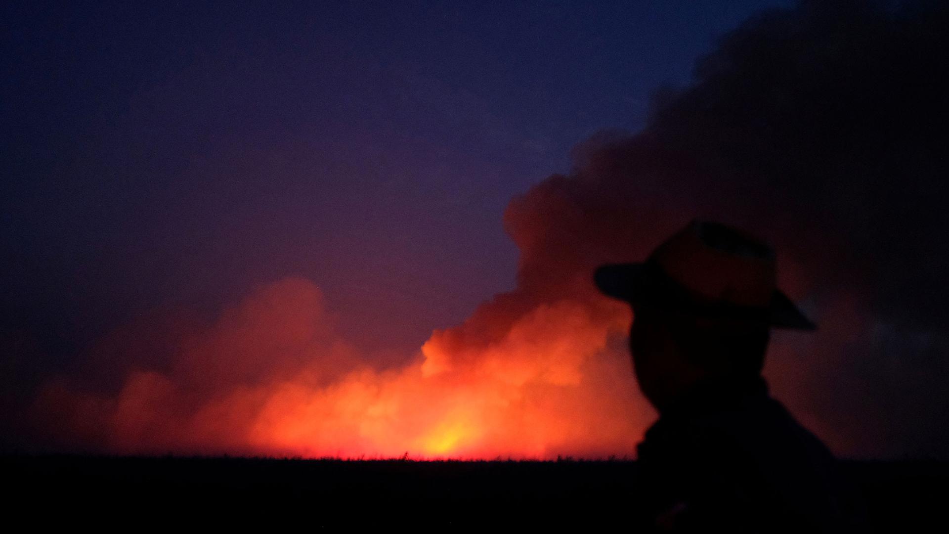 A man wearing a have is shown in the near ground, black in shadow, with a brigh orange fire seen in the distance.