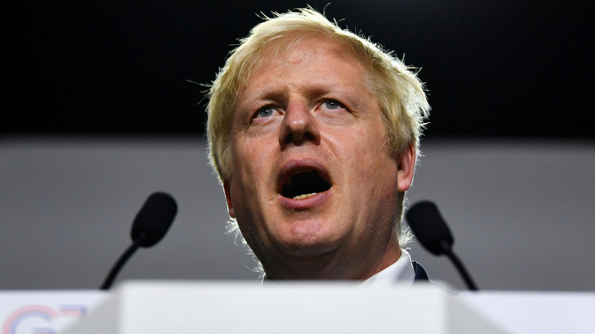 Britain's Prime Minister Boris Johnson is shown standing behind two small microphones with his mouth open speaking.