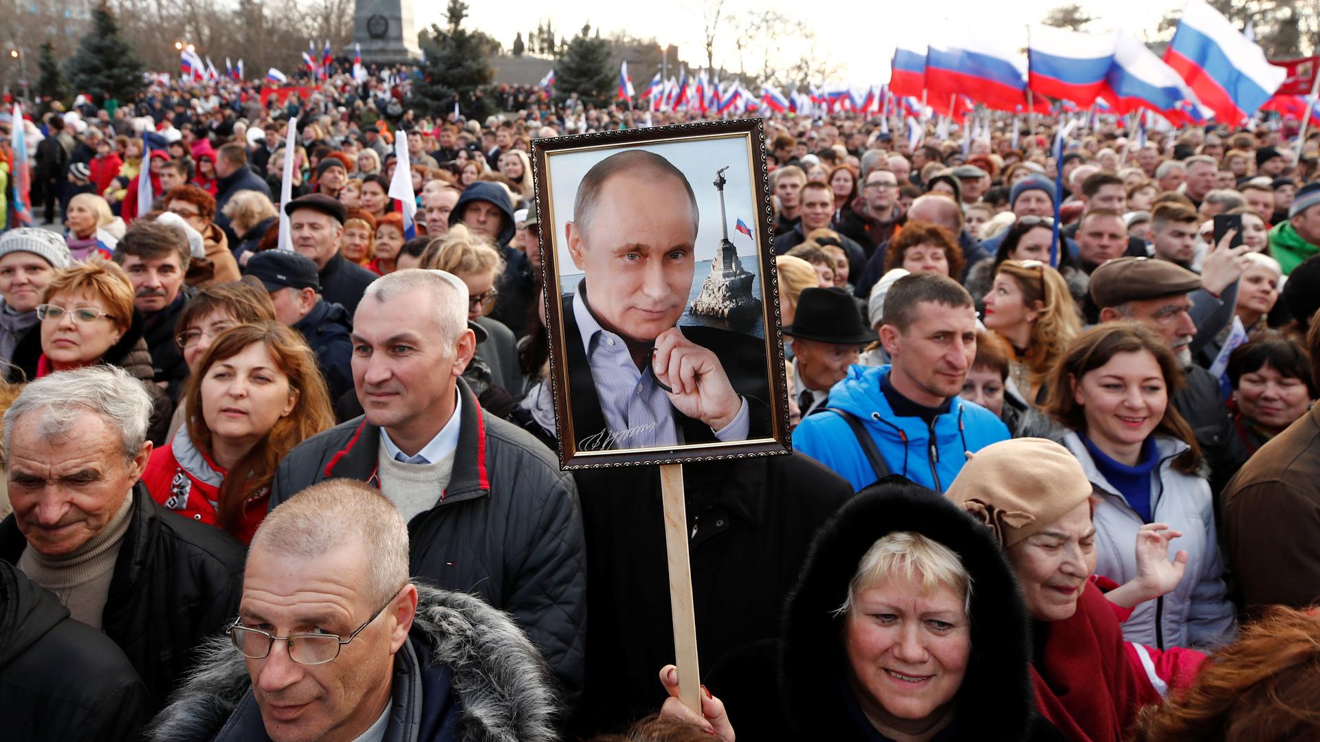 A crowd of people wave Russian flags and one holds up a photo of Putin on a sign