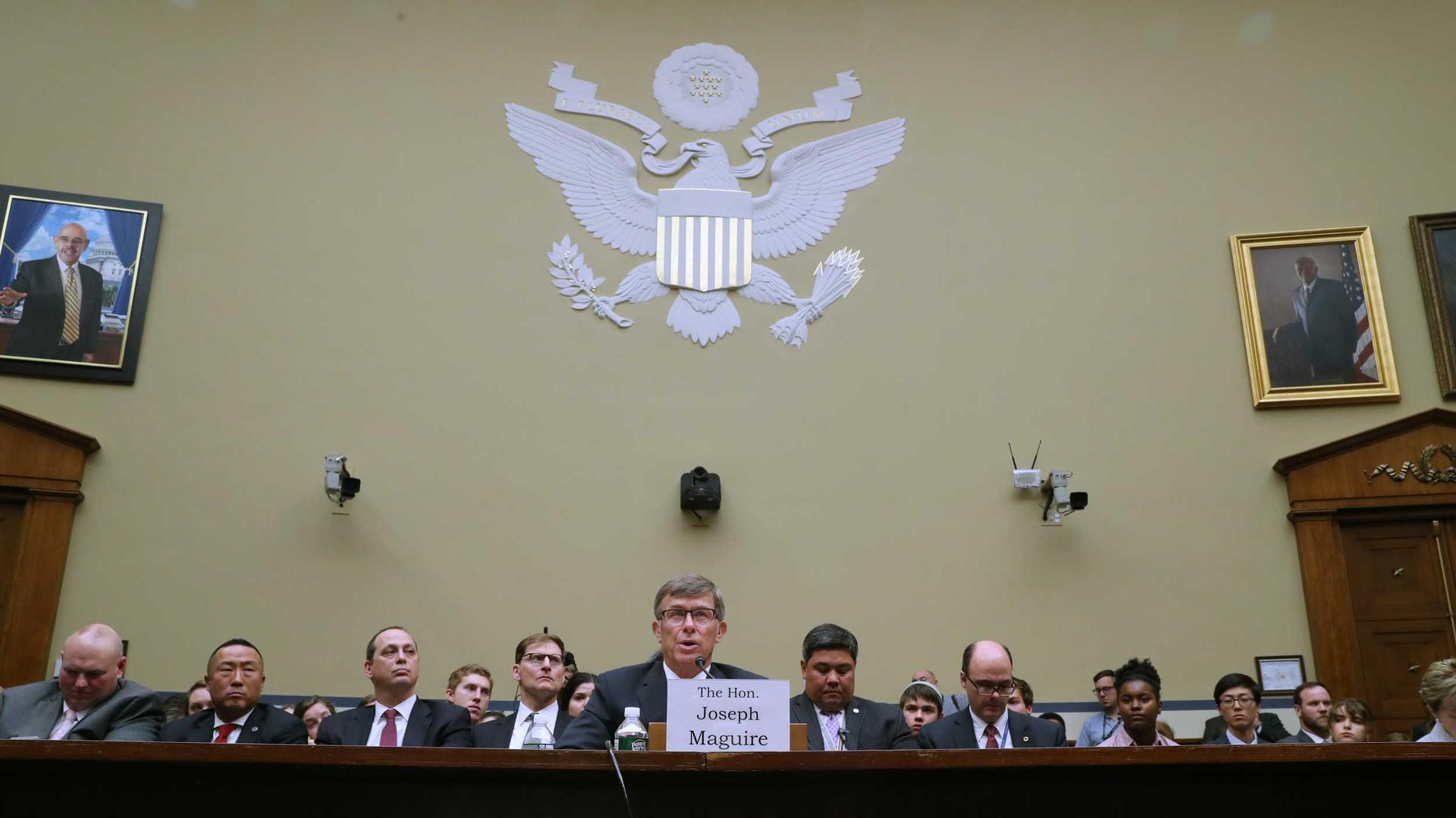 A man sits at the center of a wooden table. Behind him are more people in suits.