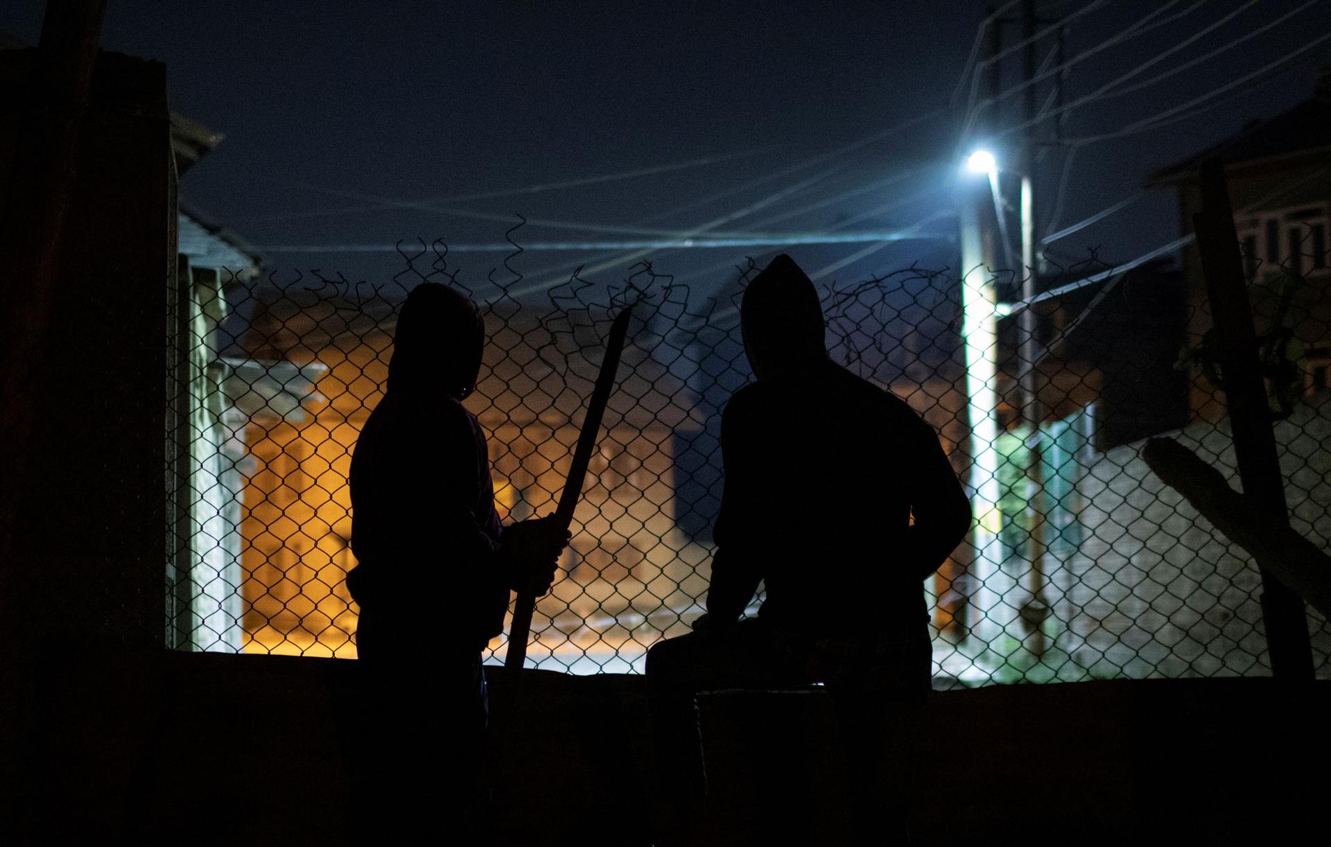 Two men are shown in shadow with a chain link fence in front of them with only the light of a streetlamp.