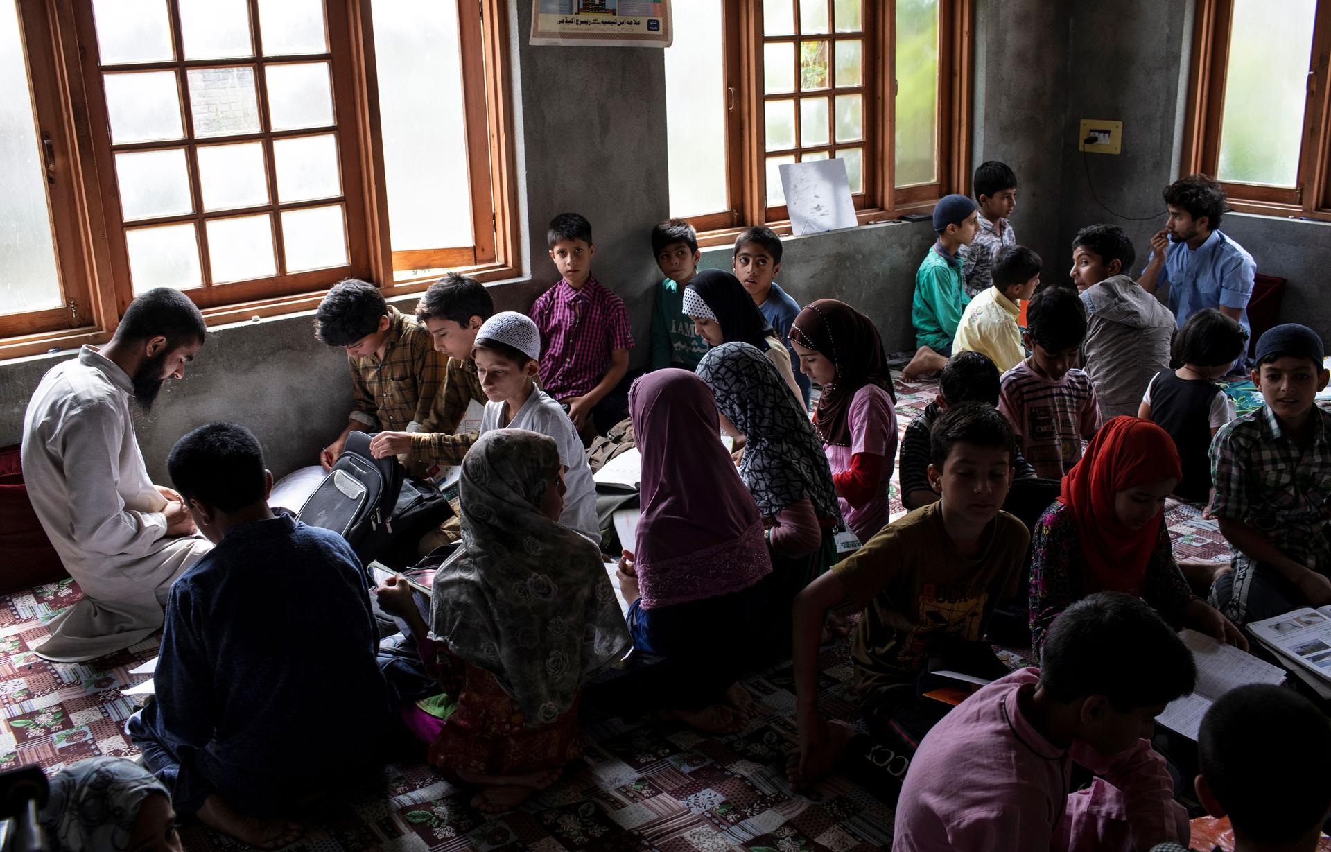 A classroom of children are shown sitting on the floor and doing school work.