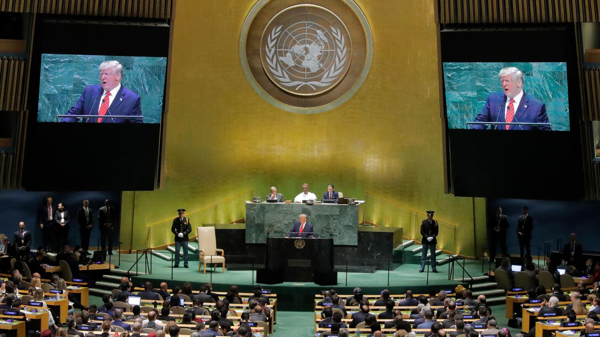 US President Donald Trump is shown on stage speaking at a black podium with live TV projections of him on either side.