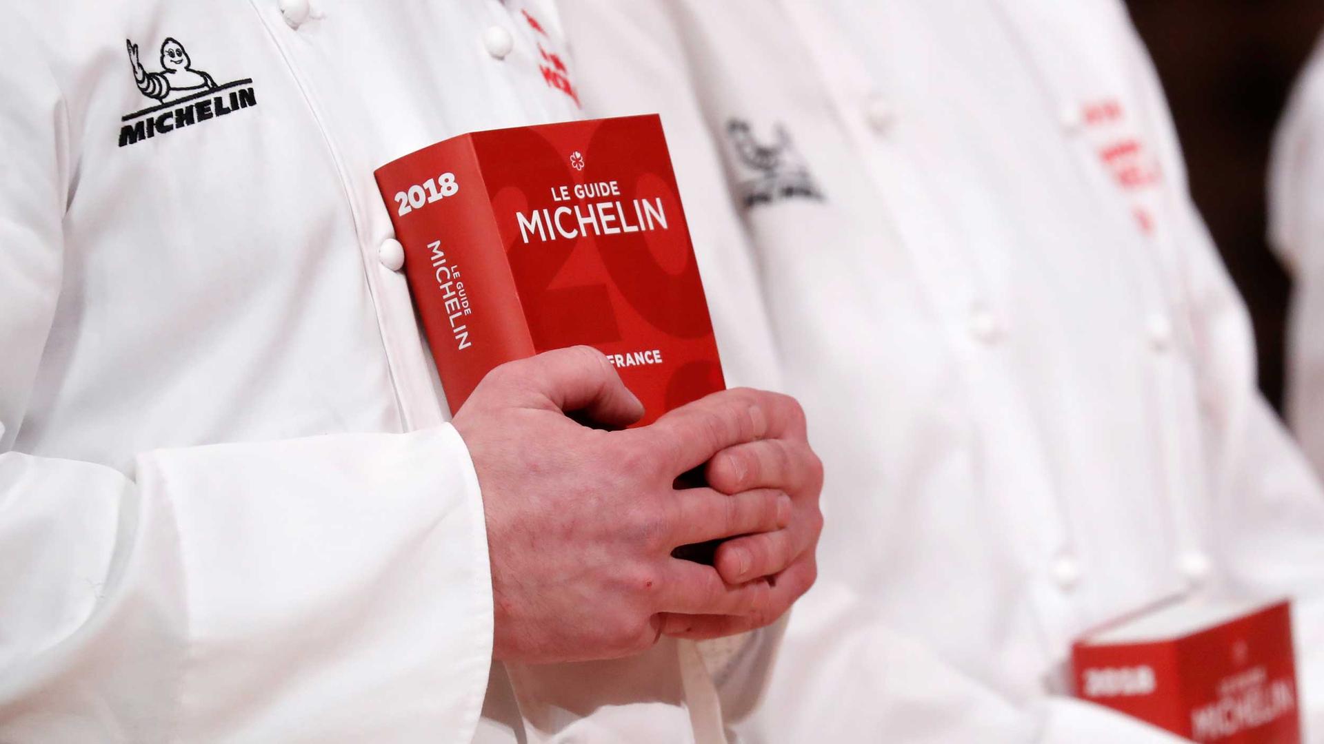A pair of hands belonging to a person wearing a white chef coat holds a red 2018 Michelin Guide.