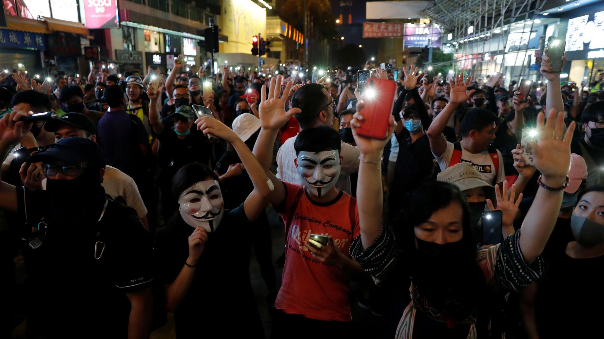 Demonstrators wearing masks hold their cell phones, with the flashlights on, over their heads as they are crowded together in a street