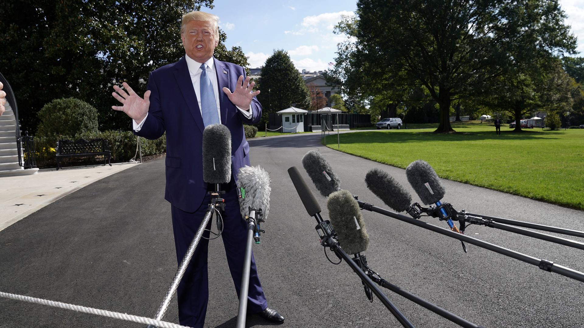 US President Donald Trump is shown standing with his hands out and wearing a blue suit with microphones in the nearground.