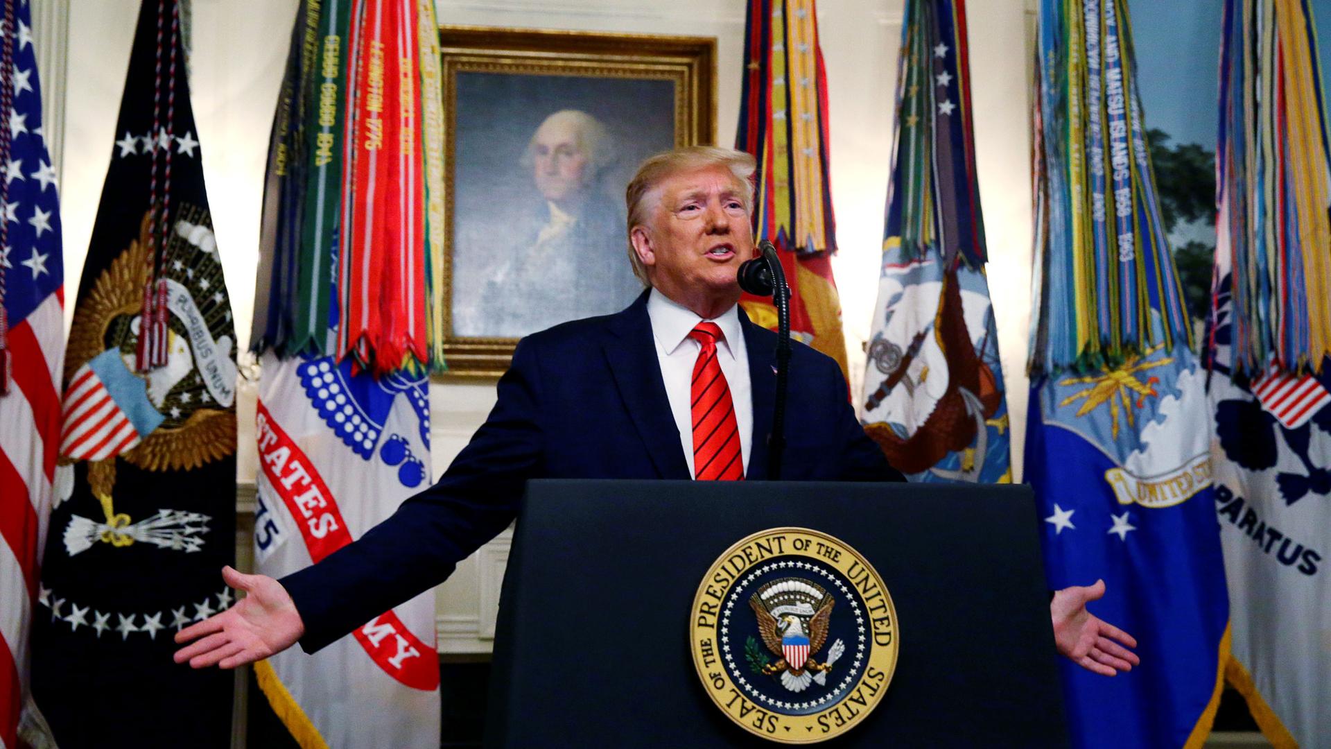 US President Donald Trump is shown standing at a podium with a seal for the US president with a painting of George Washington behind him.