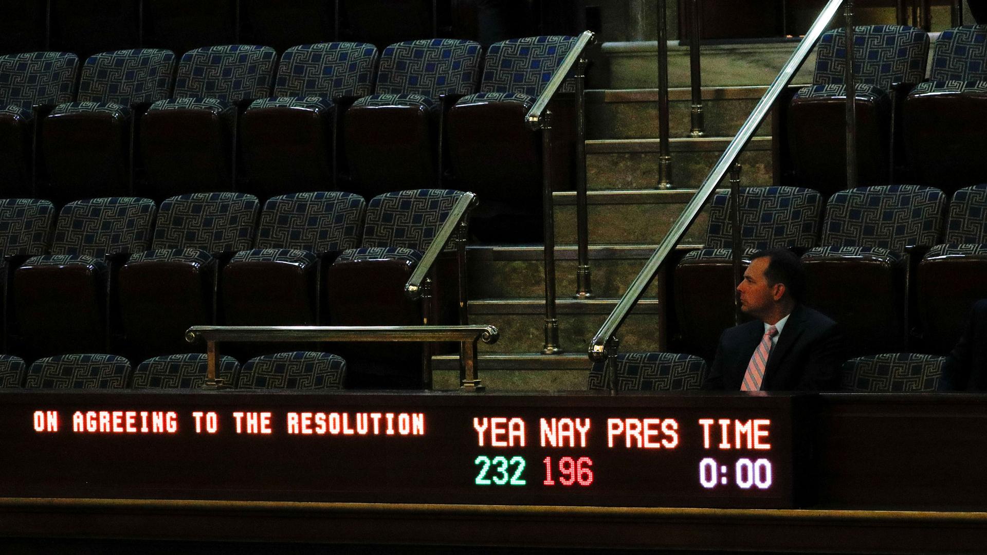 A man is shown sitting in the balcony of the US House with a vote tallying electric sign in front of him.