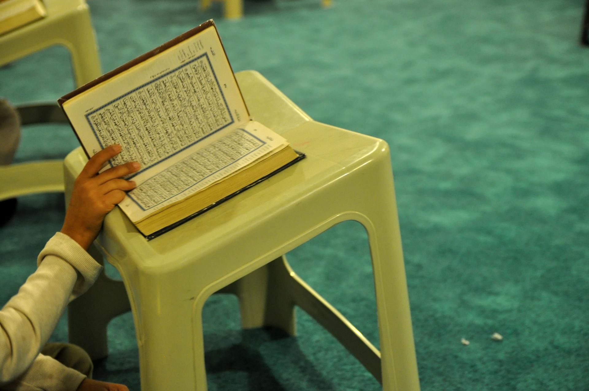 A hand holds the Quran open on a stool near green carpet