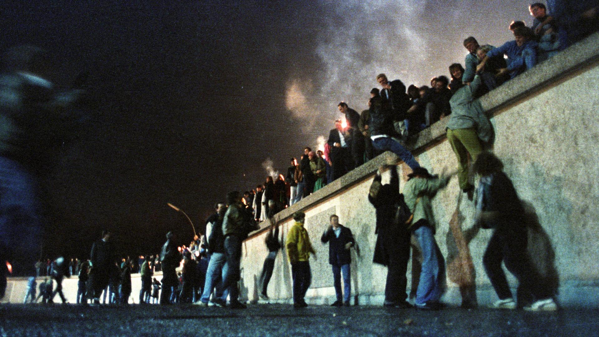 A large group of people are shown climbing on the Berlin Wall on the night it was opened.