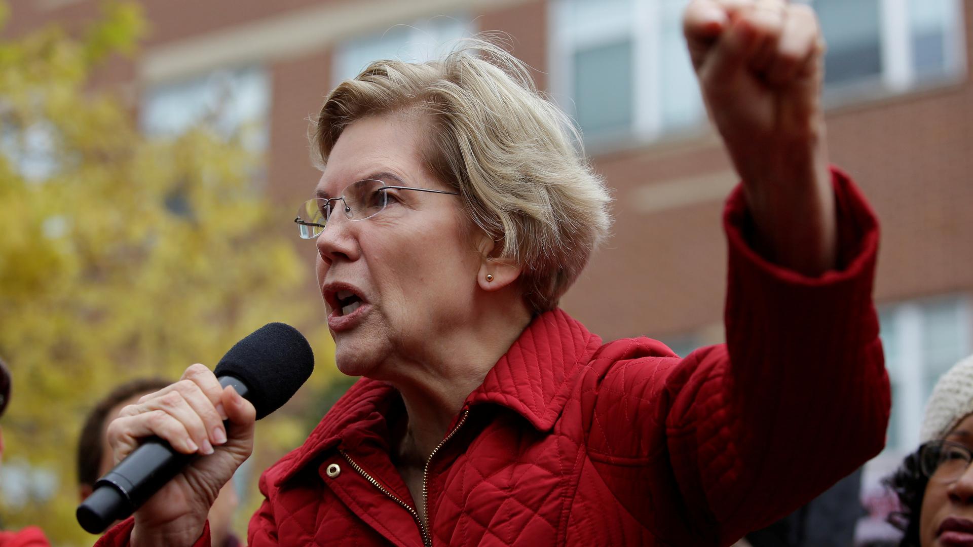 Elizabeth Warren wears a red blazer and speaks with one hand in the air.