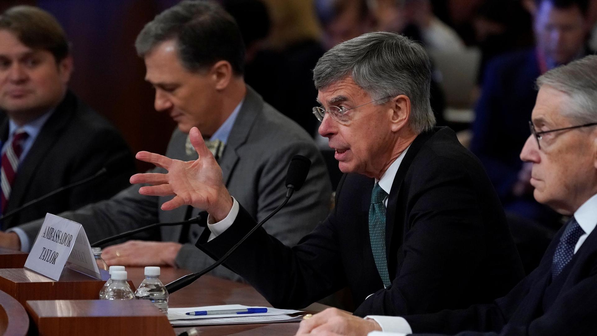 Ambassador William Taylor is shown from the side, sitting at a table with his right hand outstretched.