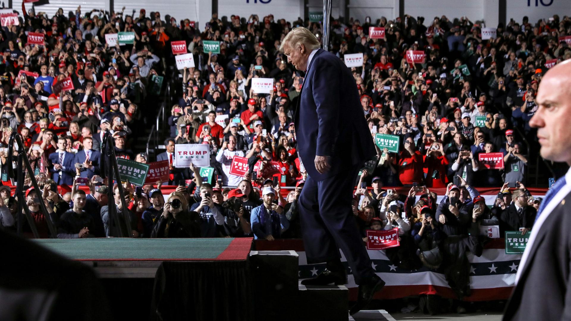 US President Donald Trump is shown walking up the steps of a stage with an audience in the distance.