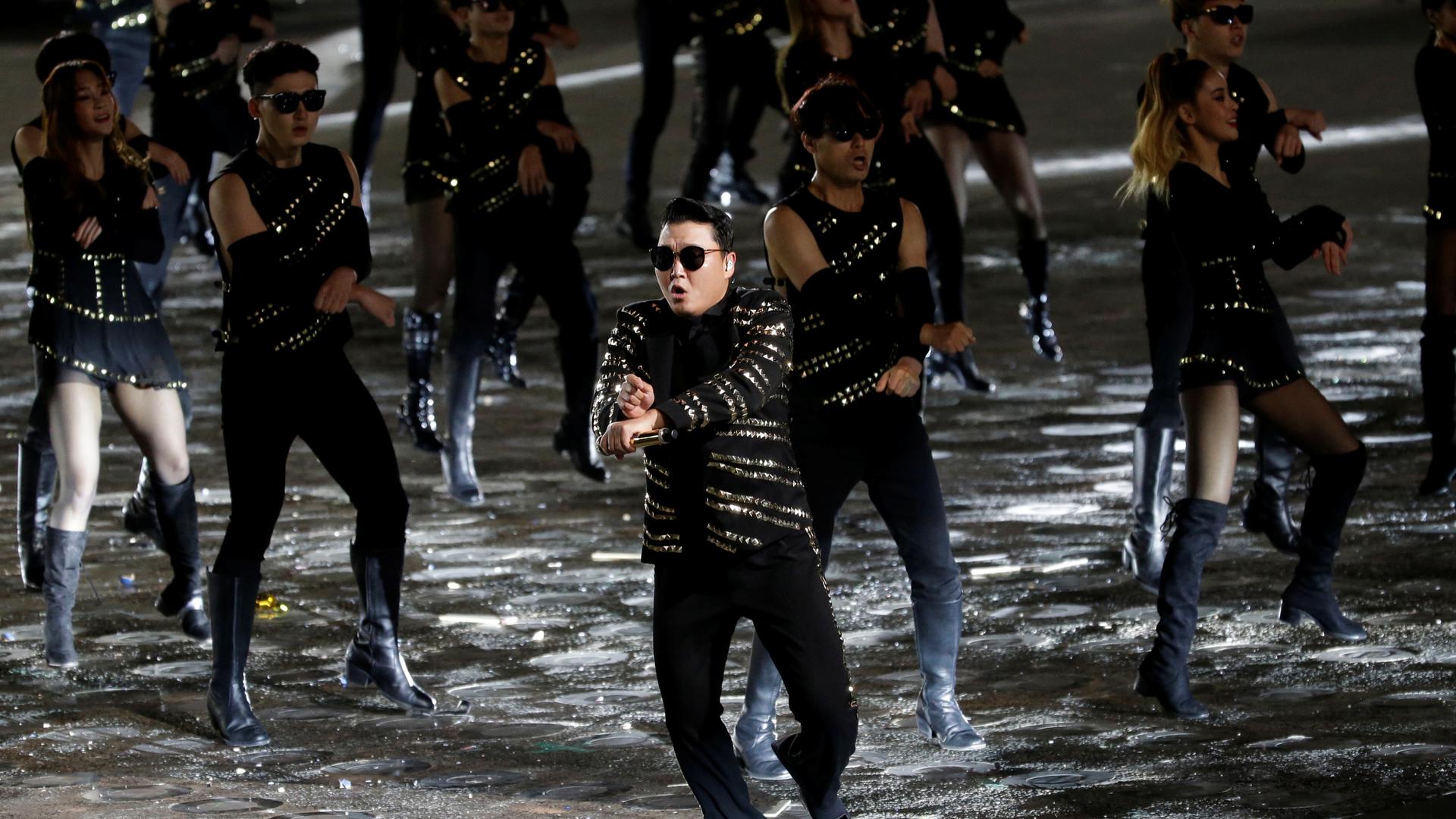 A man wears a black shiny outfit and performs with backup dancers