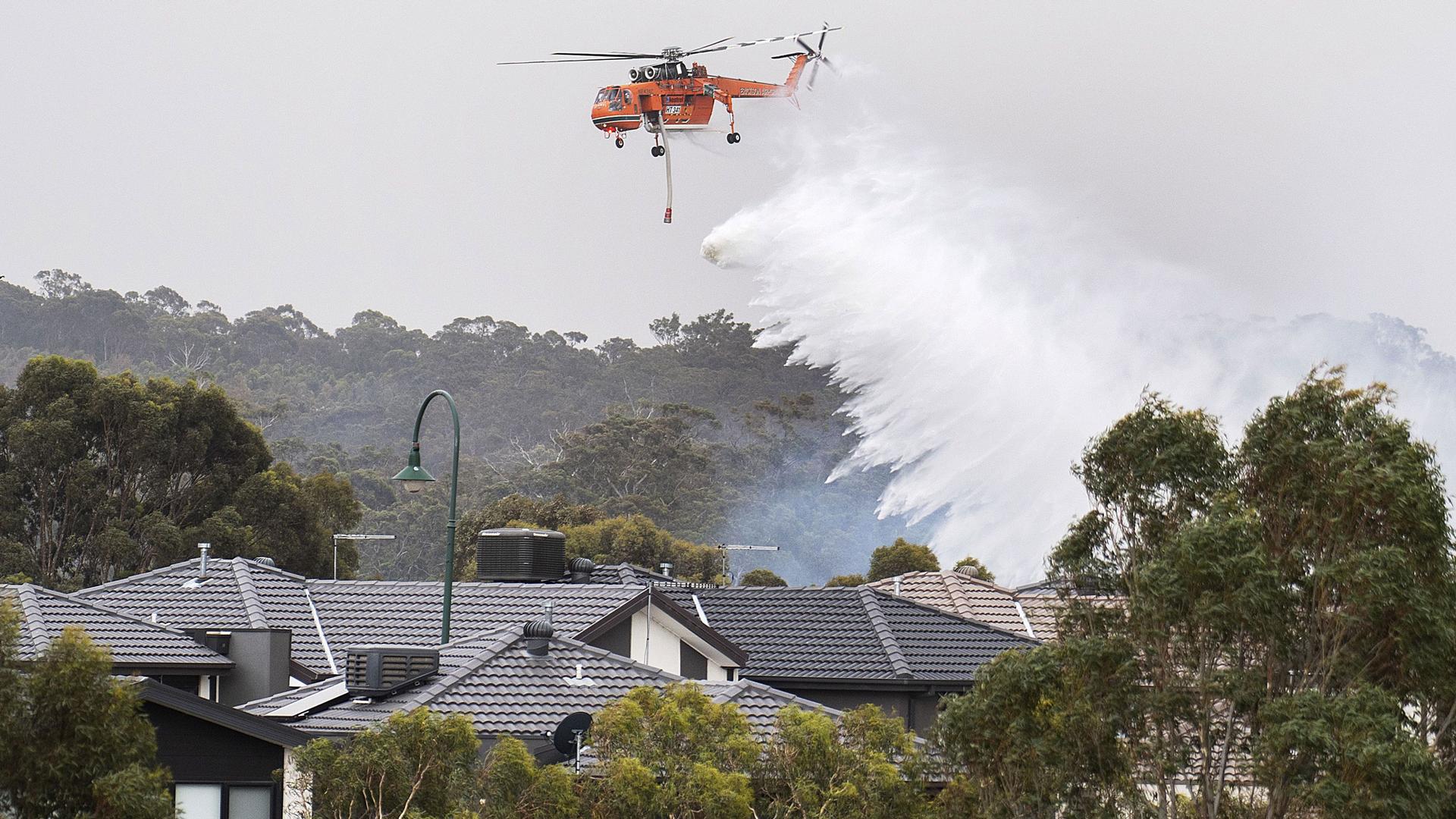 A large emergency helicopter is shown flying above several houses and dropping a large quantity of water.