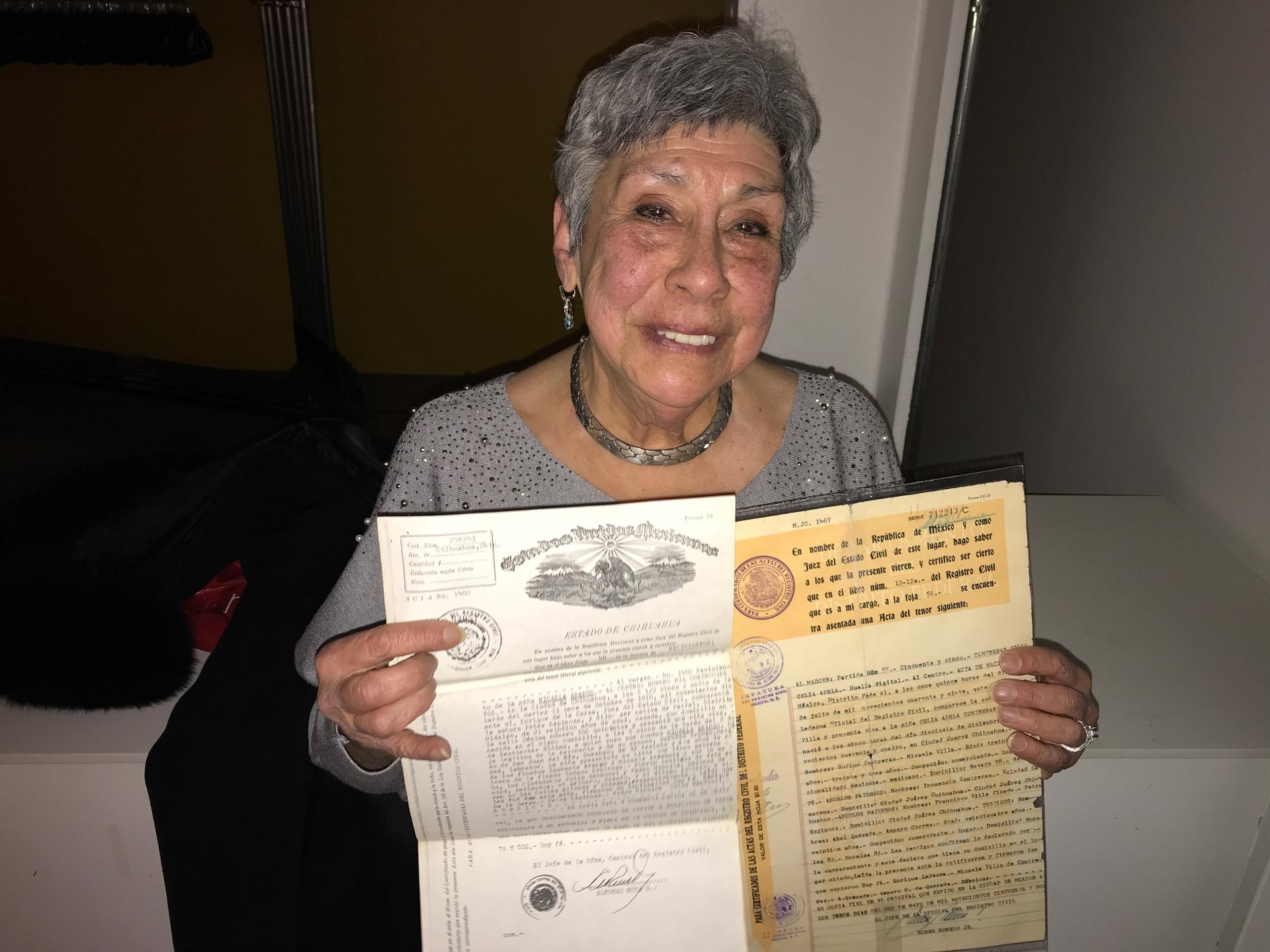 An older woman with grey, short hair and a grey sweater holds up her birth certificate