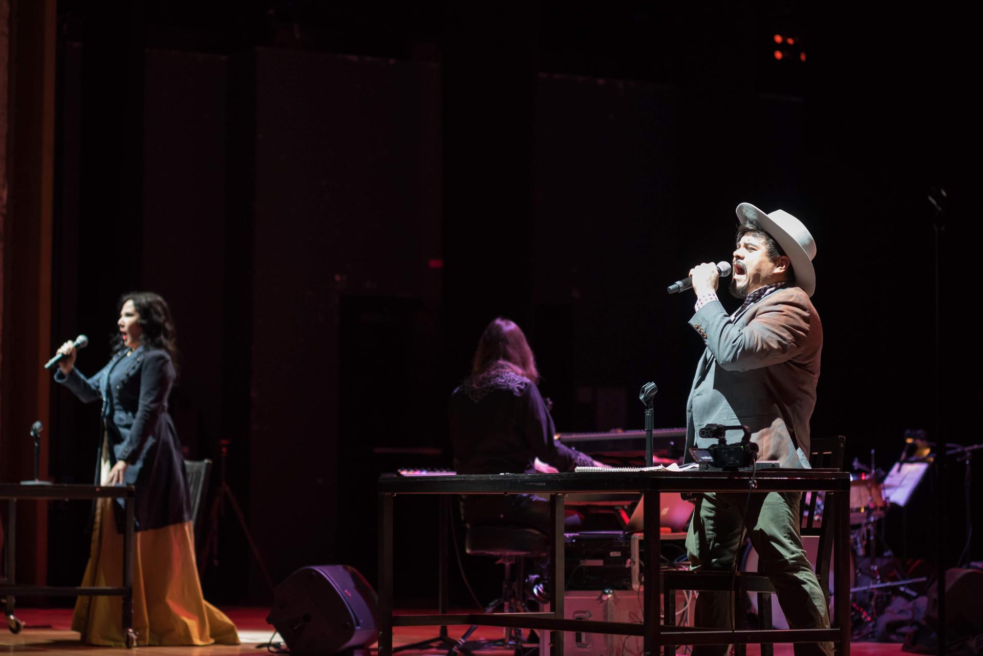 A man in the foreground dressed wearing a hat croons to the audience