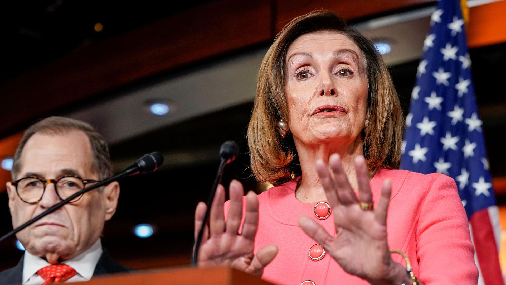 House Speaker Nancy Pelosi is shown standing at a wooden podium with microphones and several lawmakers standing behind her.