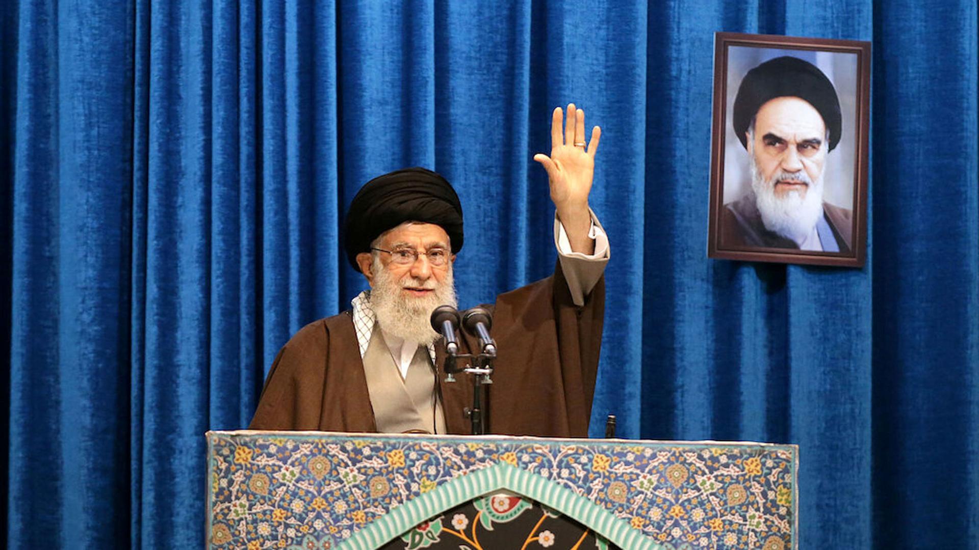 Iran's Supreme Leader Ayatollah Ali Khamenei his shown standing at a podium with his hand raised.